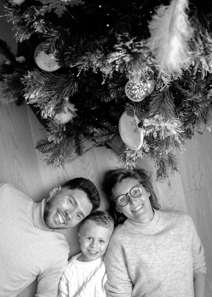 Un couple et leur enfant allongés sur le sol, souriant, pris en photo du dessus, devant un arbre de noël