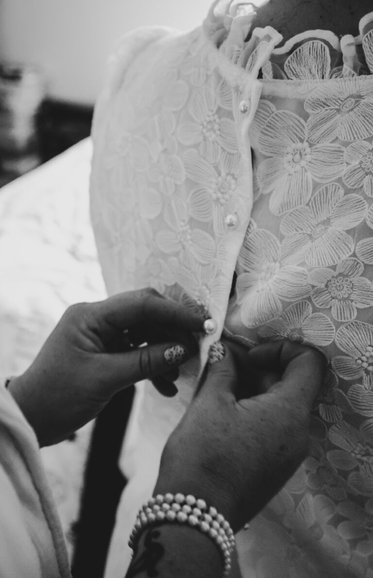 Shooting photo mariage - gros plan sur le dos de la robe de la mariée en dentelle, une main ferme délicatement les boutons